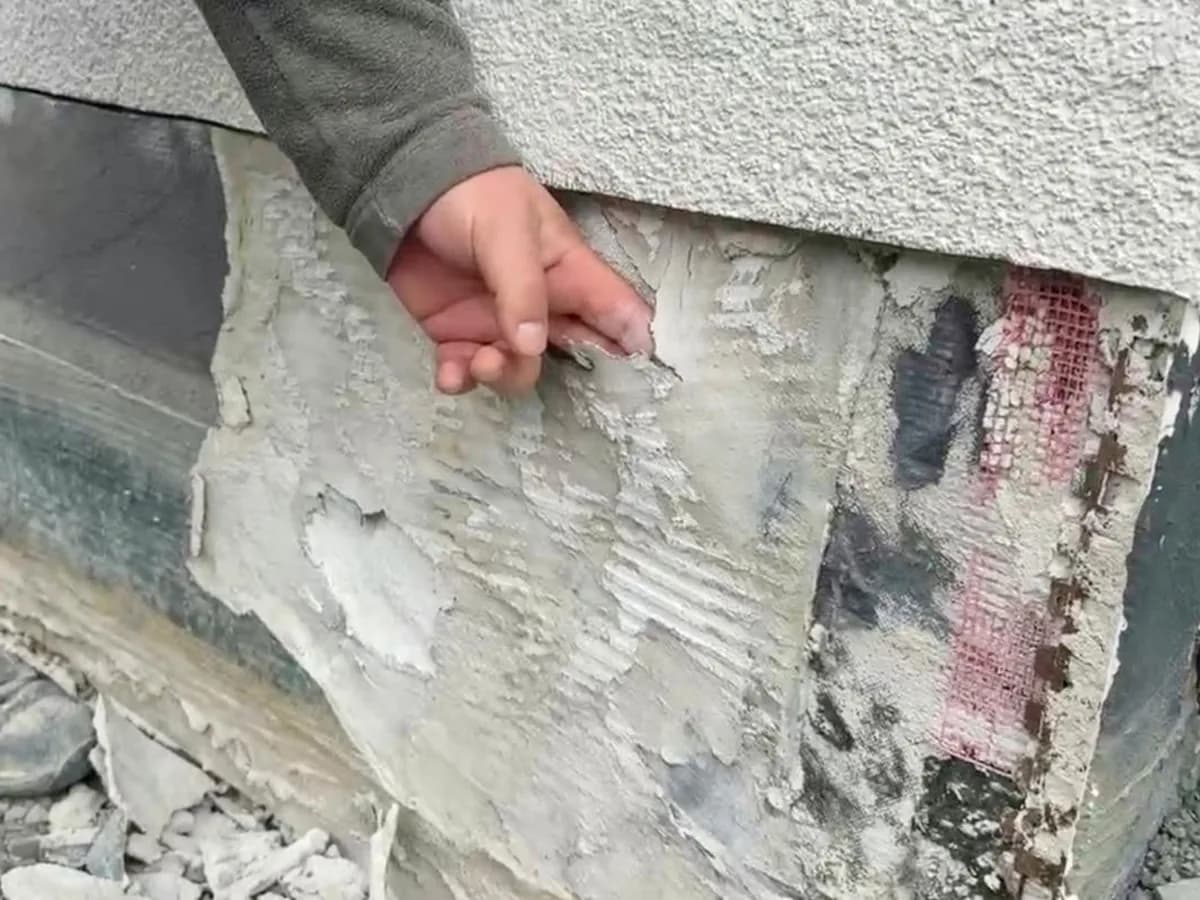 Sockelbereich einer Hausfassade: Die Abdichtungsschicht löst sich flächig von der Wand und wird von Hand abgezogen, darunter kommt das Armierungsgewebe zum Vorschein